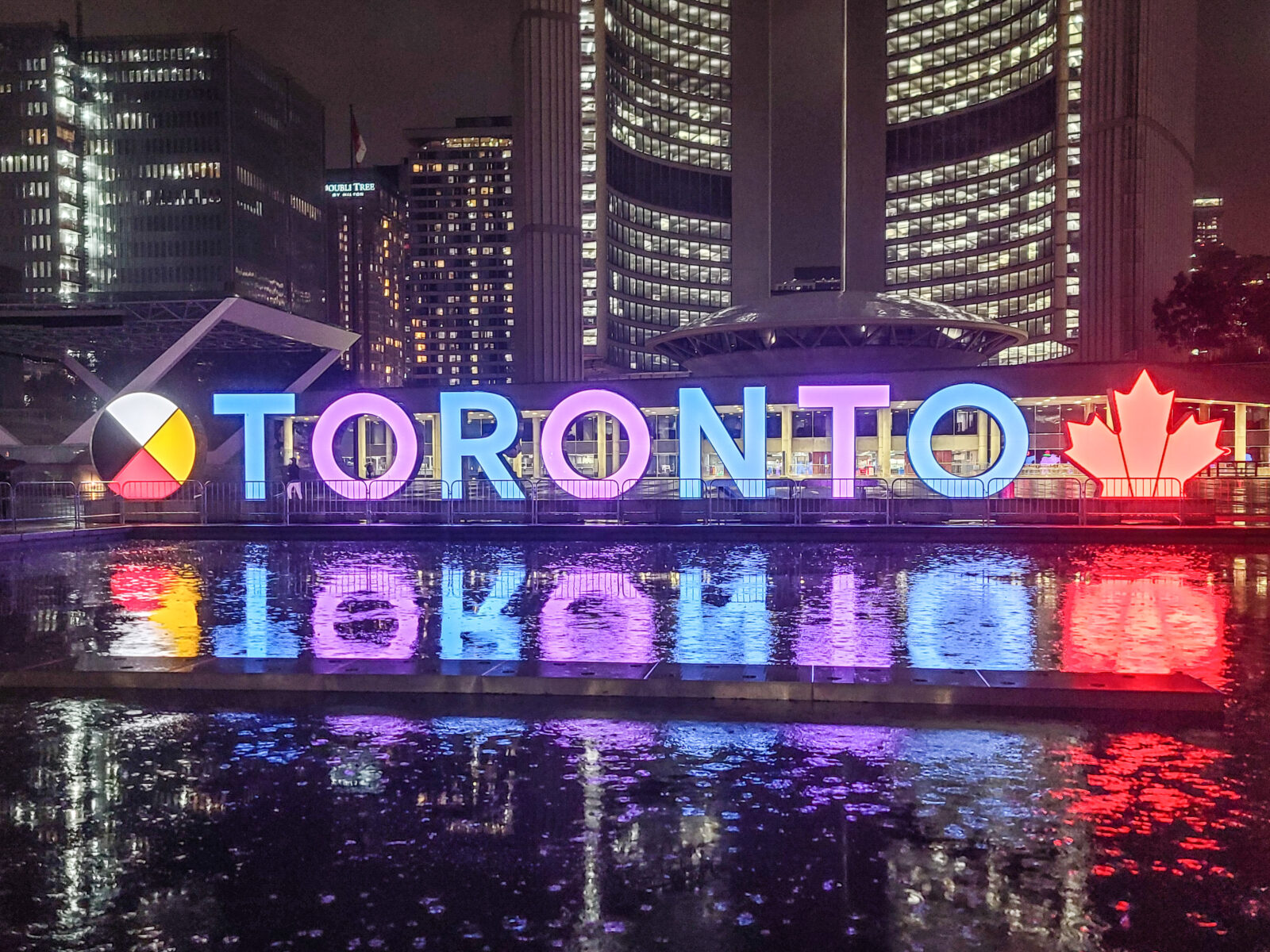 3D Toronto Sign lit purple and blue, reflecting in the pool, with City Hall towers in the background