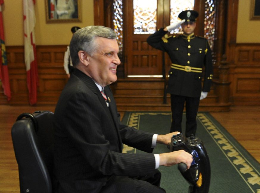 A smiling David Onley passes by the camera on his mobility scooter inside Queen's Park, in profile and smiling. In the background, a guard is saluting.