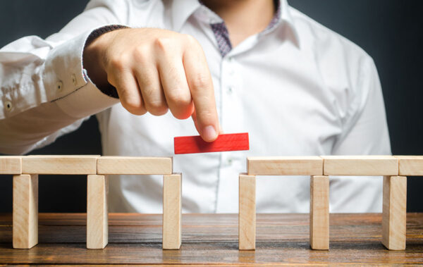 A man is holding a red wooden block between his fingers and placing it into position to complete a bridge made of wooden blocks that aren't painted.