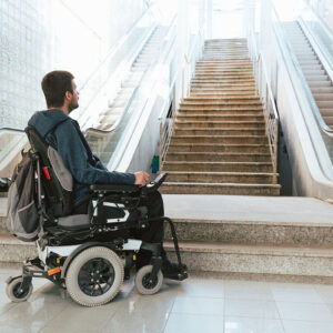 A man is in a power wheelchair at the base of stairs and escalators. He's looking up the escalator. There's no ramp, so the stairs and escalators aren't accessible.