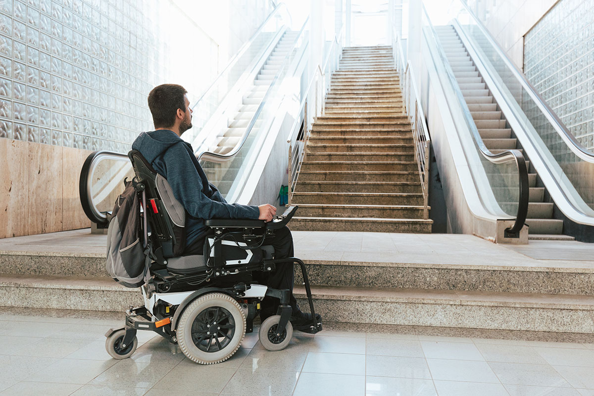 A man is in a power wheelchair at the base of stairs and escalators. He's looking up the escalator. There's no ramp, so the stairs and escalators aren't accessible.