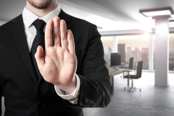 A businessman is holding up his hand in a stop gesture. In the blurred background, there are desks and work tables in an open-office environment.