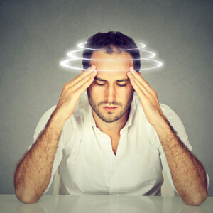 A man is sitting with his eyes closed and his elbows resting on a table. His arms are raised and his fingertips are pressing against his temples. There are swirls around his head, representing dizziness.