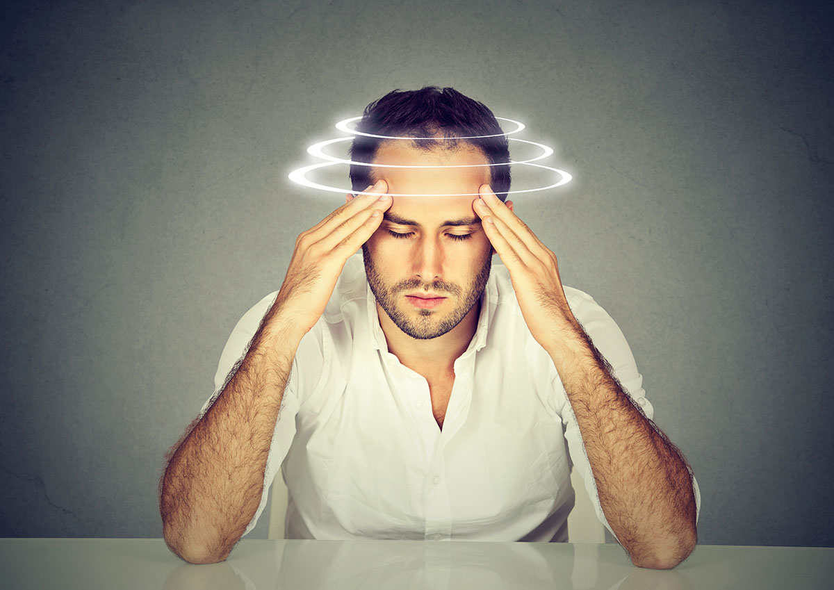 A man is sitting with his eyes closed and his elbows resting on a table. His arms are raised and his fingertips are pressing against his temples. There are swirls around his head, representing dizziness.