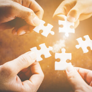 Five hands holding puzzle pieces, bringing them together.