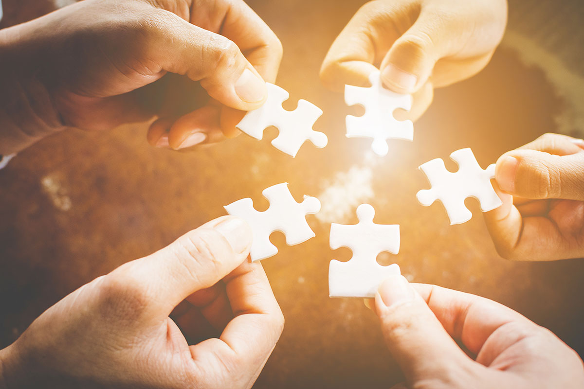 Five hands holding puzzle pieces, bringing them together.