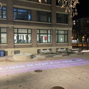 A Light It Up! For NDEAM National Disability Employment Awareness Month purple light banner shines on the sidewalks of Elgin Plaza at Red River College Polytechnic in Winnipeg on October 19, 2023. It's shining in front of a campus building.